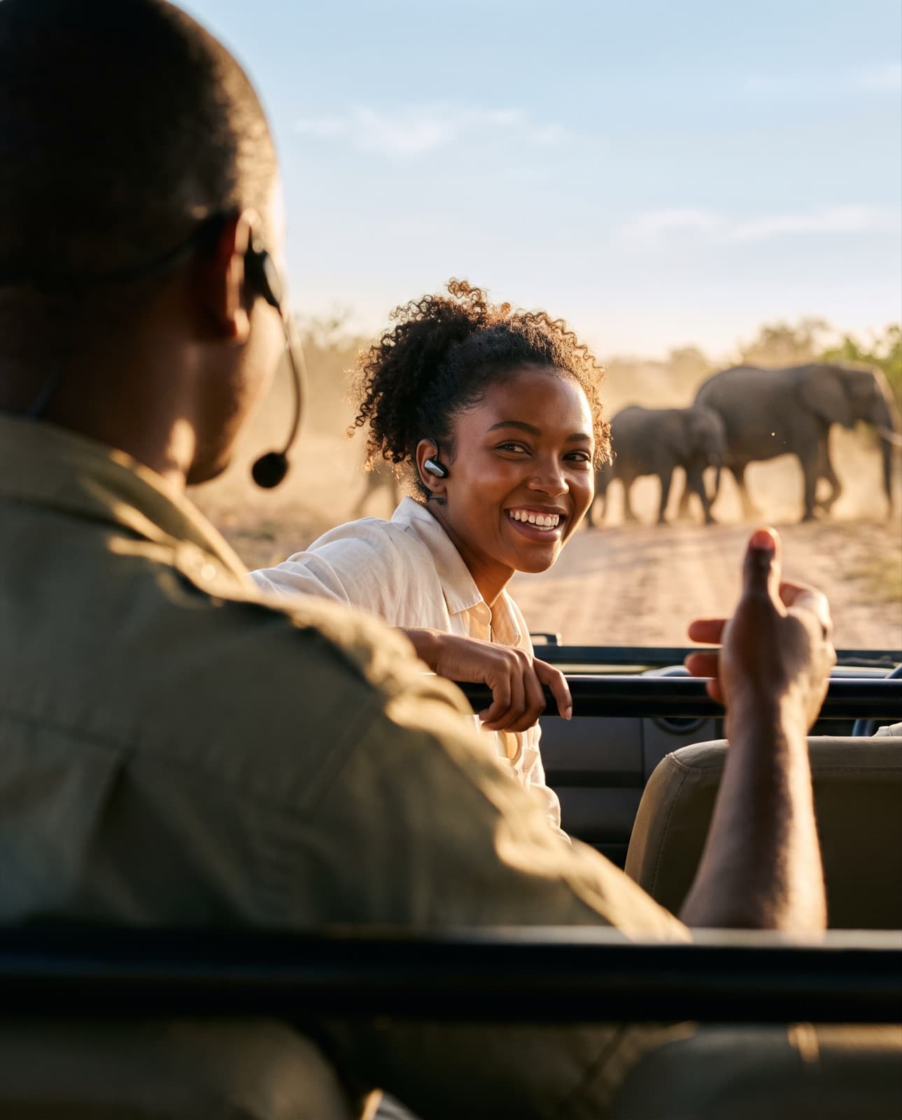 A smiling guest in an open safari vehicle, wearing a small earpiece, listens to her ranger's headset microphone as elephants cross the track behind her